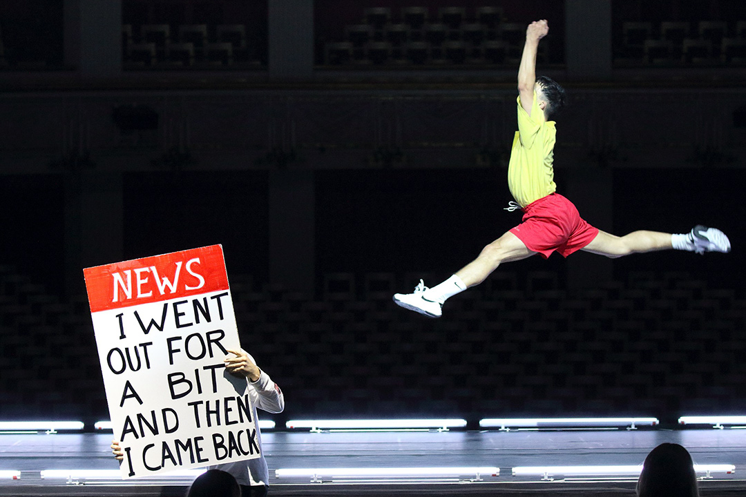 Performer leaps on stage with news headline sign.