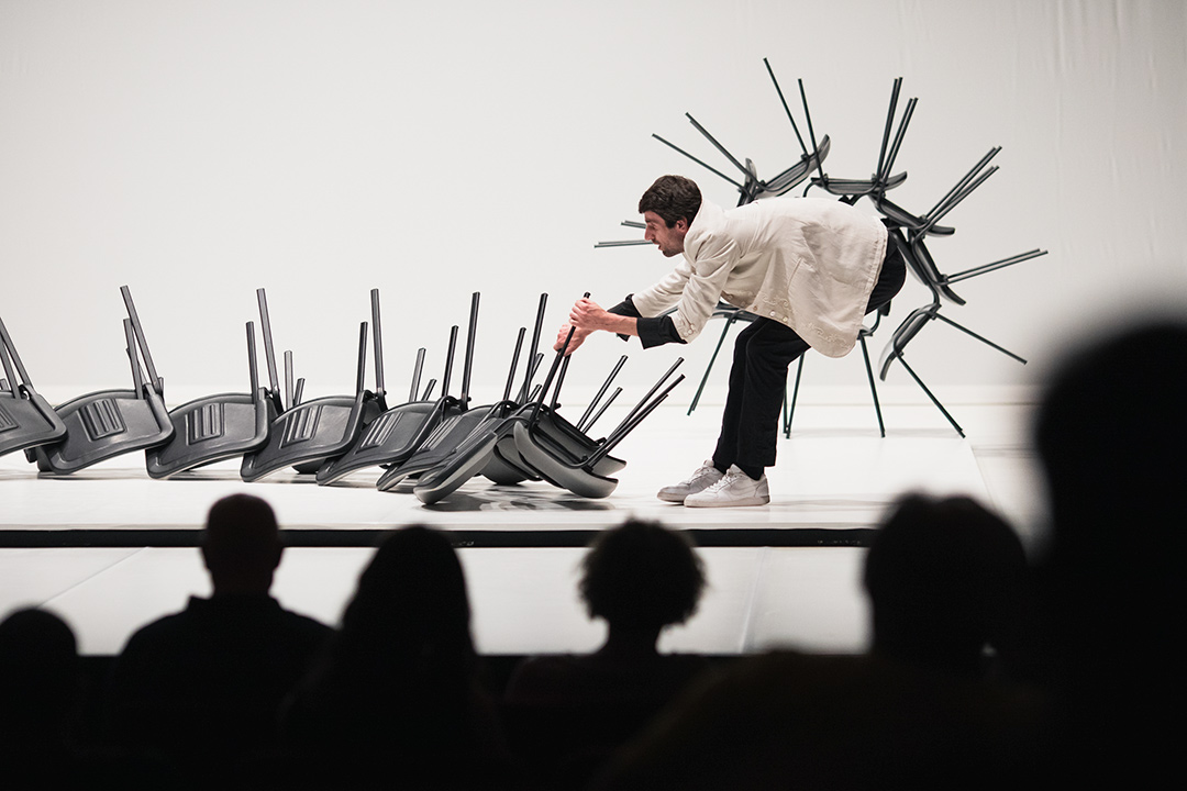 Man performing with stacked black chairs on stage.