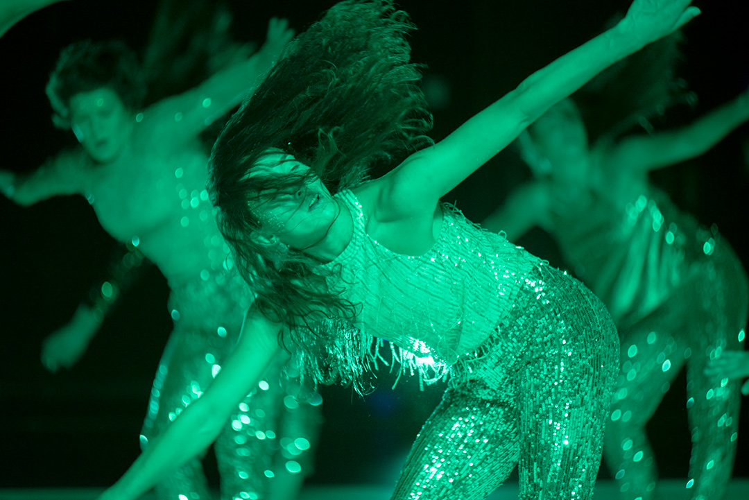 Dancers performing in green lighting, wearing sequined costumes.