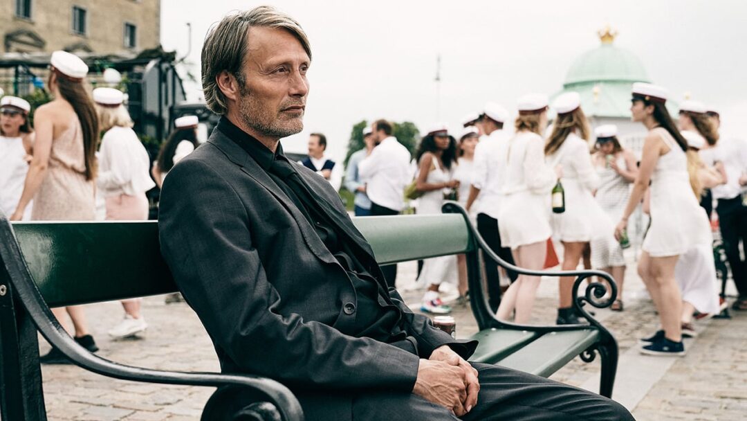 Mads Mikkelsen se sienta en un banco mientras los estudiantes festejan detrás de él en la escena final de la película Another Round de Thomas Vinterberg