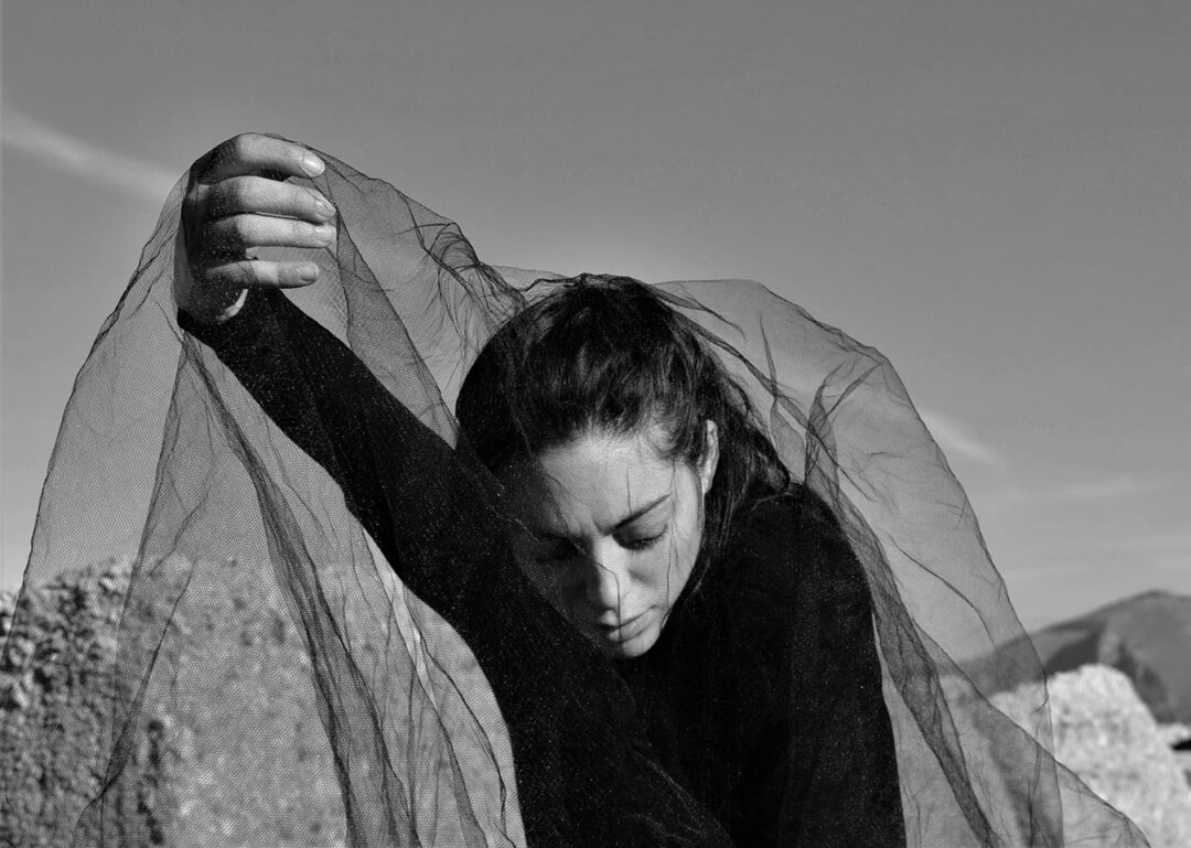 Black and white portrait of a woman's upper body, face looking down and one arm extended forward holding a net mesh that cloaks her. The background is of stony hills and sky