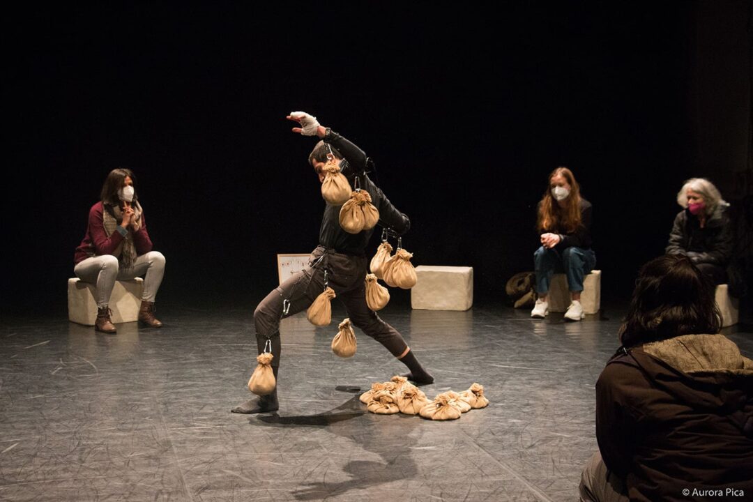 Man with sandbags tied to his body in the centre of a circle of stone blocks on which sit various audience members in covid masks