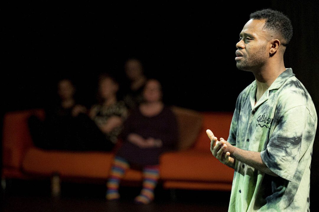 Black man in green shirt at right of photo, holds arms together, one amputated at the wrist. Behind on a sofa are the blurry images of other participants