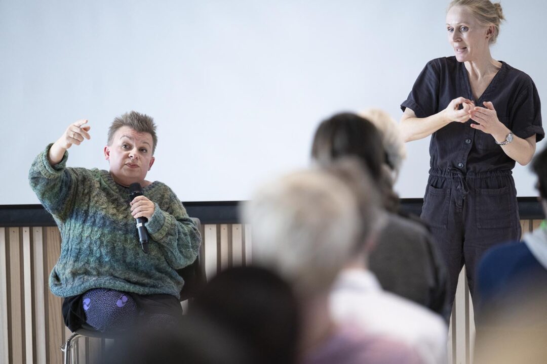 Seated woman with raised arm speaks into microphone. A woman interprets using sign language.In front, the backs of heads in the audience