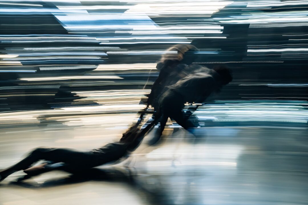 Motion blur photo of two dancers dragging a third one on her back at speed across a stage, with blurred lighting in the background