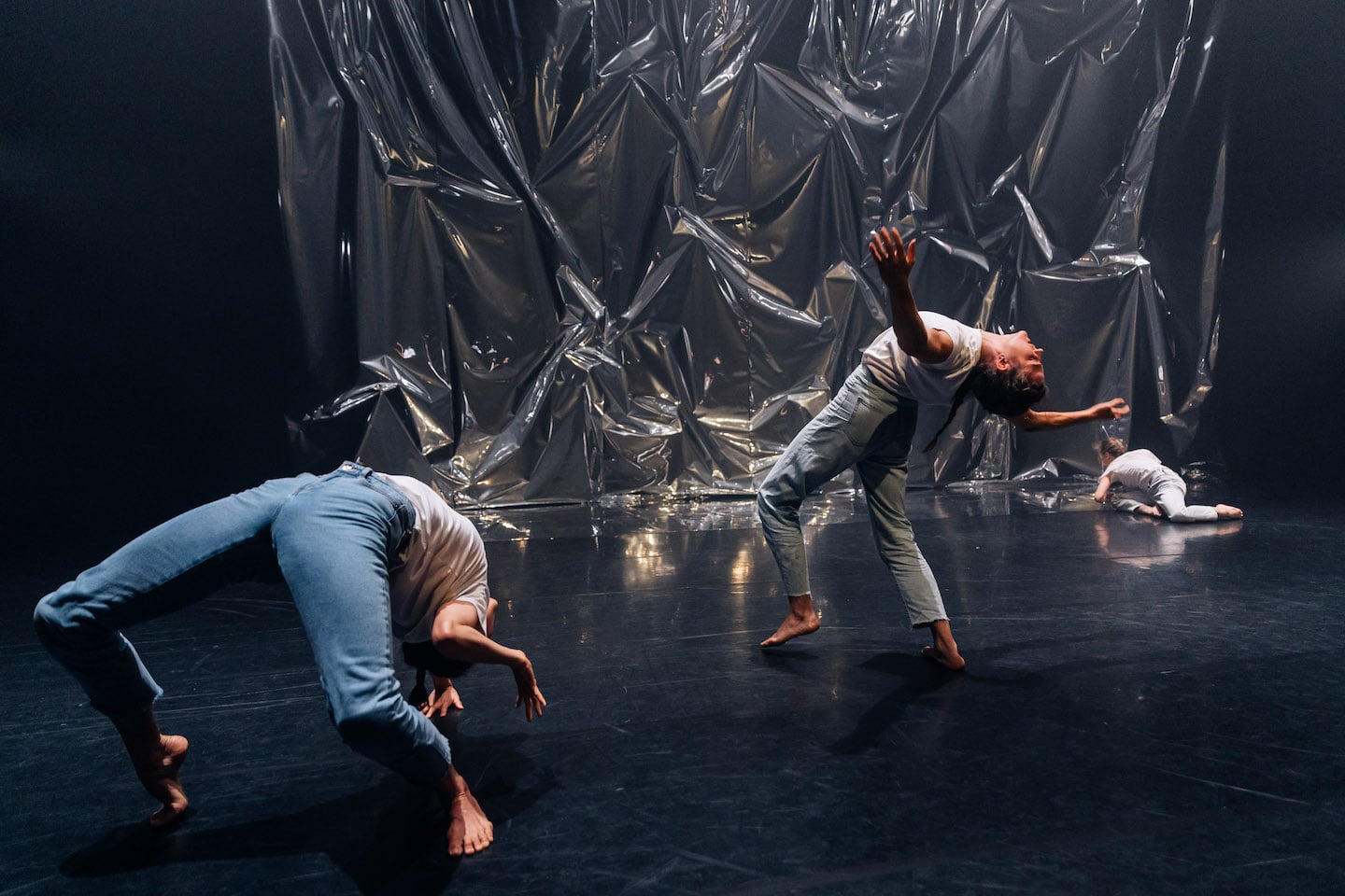 Three women in jeans and t-shirts on a stage backed by crinkled plastic curtain. Two front women in deep, unbalanced back bends. The woman at the back is on the floor already.