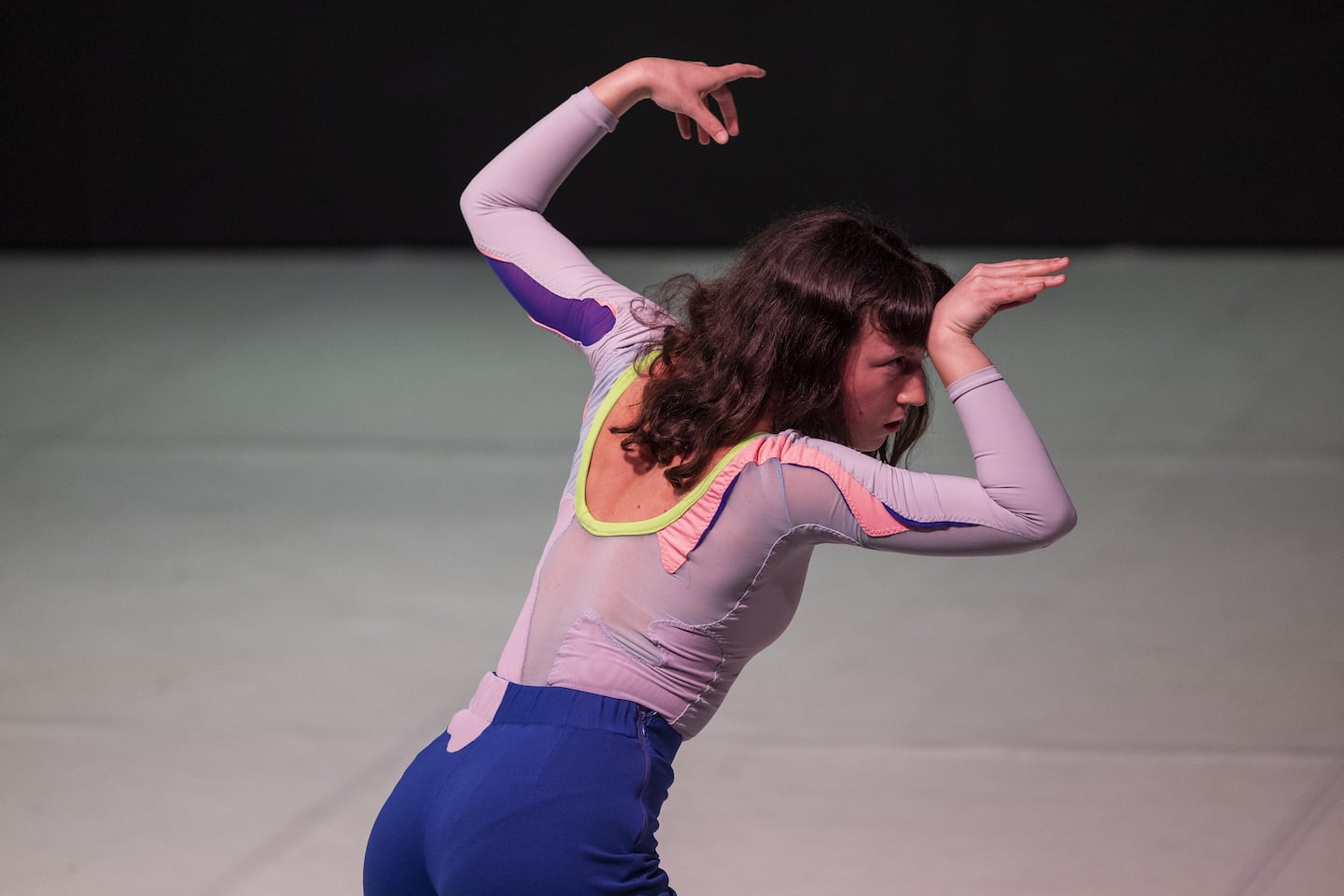 A woman in light purple top and dark blue trousers, patched with a coral pattern, looks intently to the right, arms bent and hands angled like antlers