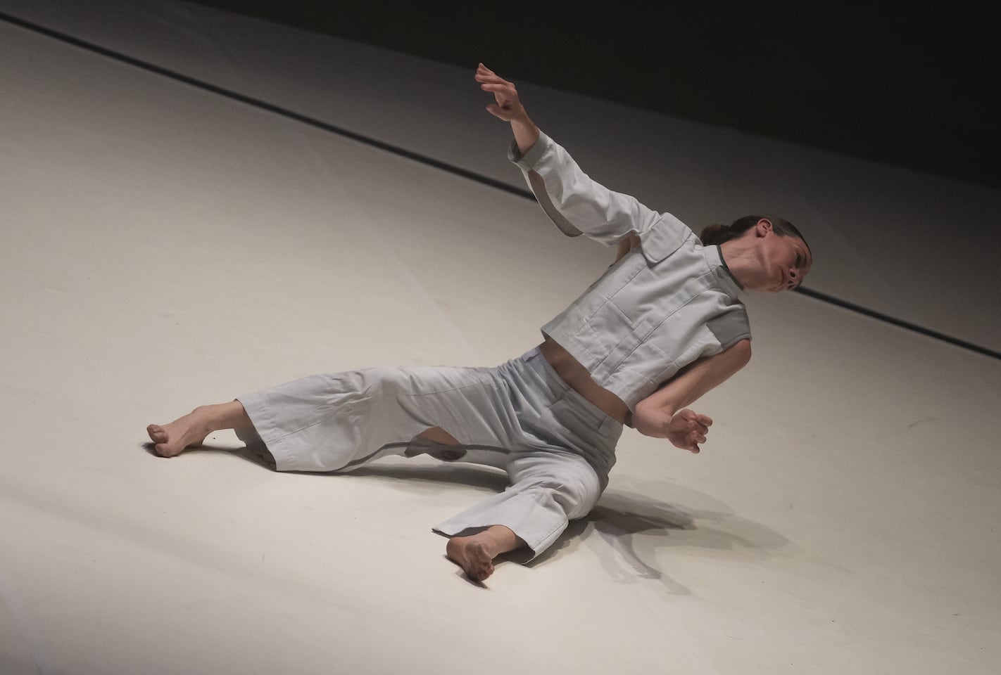 A woman in light off-hite tunic and trousers is sitting on the floor, leaning backwards almost to the point of overbalance, head tilted far away from us.