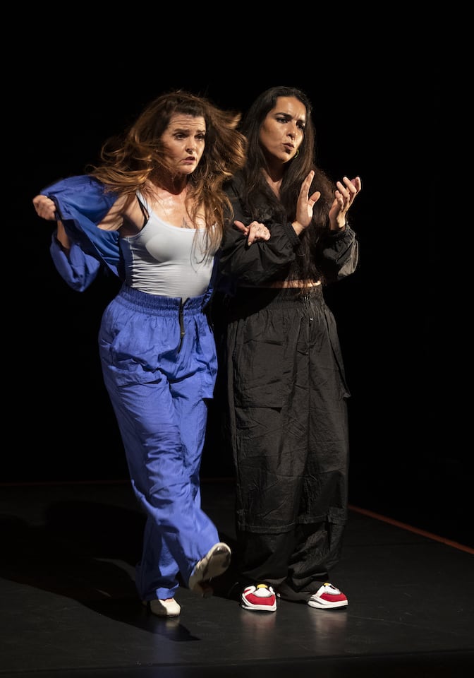 Two women stand side by side, arms linked. One in dark boiler suit is clapping and singing. The other in bright blue is in mid-step, training top half open to reveal sports top beneath.