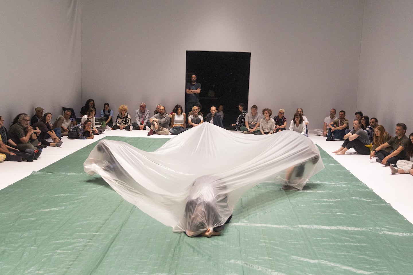 A white room with spectators sitting round the edge The middle is covered with green plastic flooring, on which four figures crouch in an outward-facing diamond, stretching the corners of a large plastic sheet as if they were the corners of a marquee or tent
