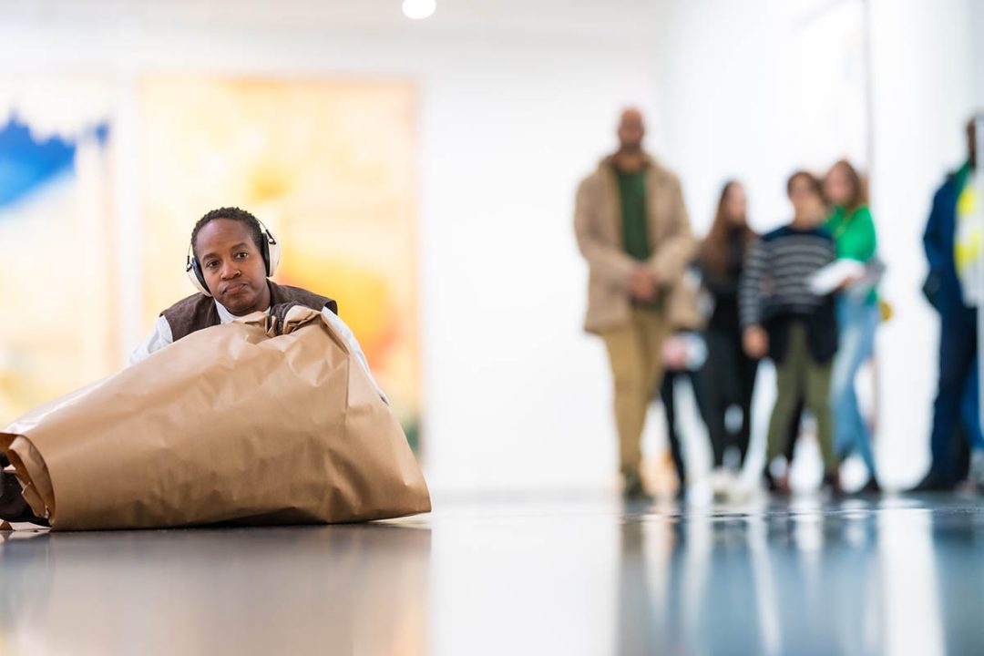 Person listening to headphones in art gallery, with a large cone of brown paper in front.