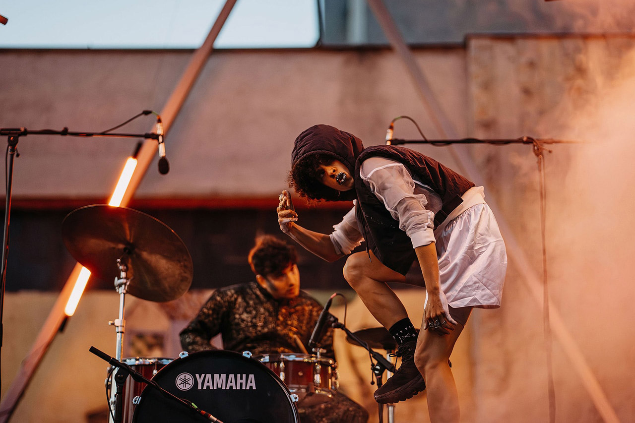 Performers on outdoor stage with microphones and drums.