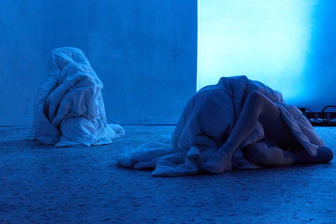 Crumpled piles of blue-lit sheets, one concealing the body of a dancer