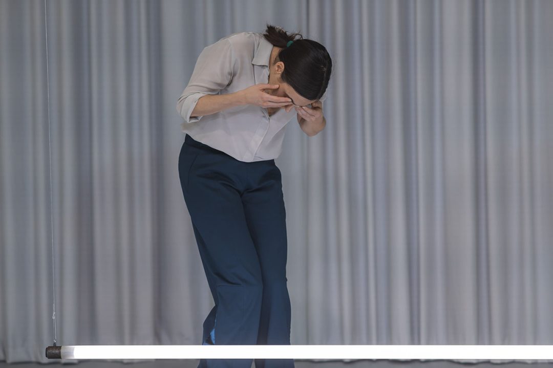 Woman in blue trousers and white shirt, leaning over with hands over eyes, above fluorescent strip light
