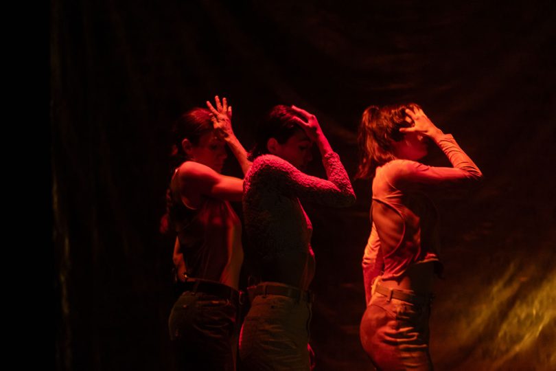 Three performers move in unison beneath dramatic red lighting. Their expressive poses create a striking, atmospheric stage moment.