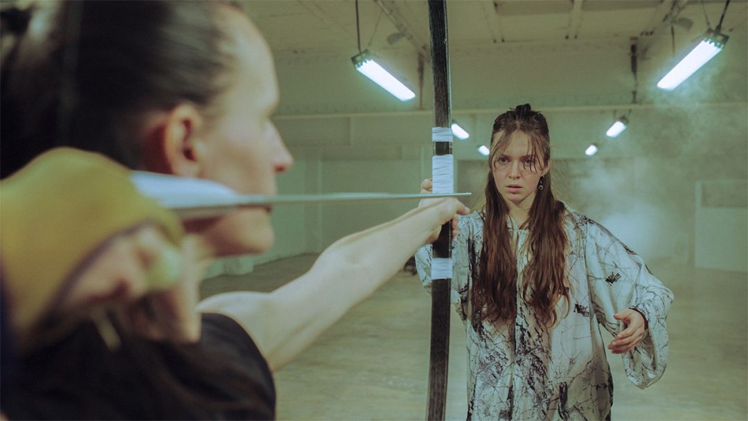 A tense standoff unfolds in a stark, empty room. One woman holds an arrow inches from the other, the air thick with suspense.