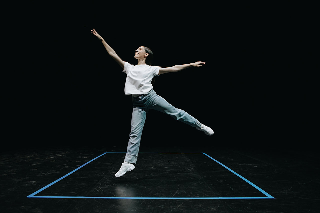 A dancer moves gracefully within a striking blue square on a dark stage. Her poised leap captures a moment of balance and freedom.
