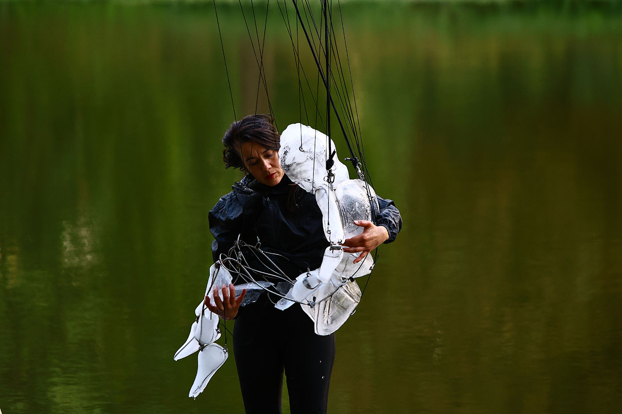 A performer cradles a suspended marionette against a tranquil lakeside backdrop. The scene blends art, movement and reflection.
