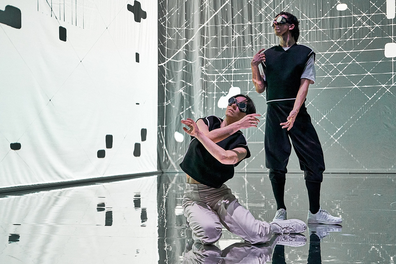 Two performers move through a striking, futuristic stage environment. Digital projections and reflective flooring enhance the dramatic atmosphere.