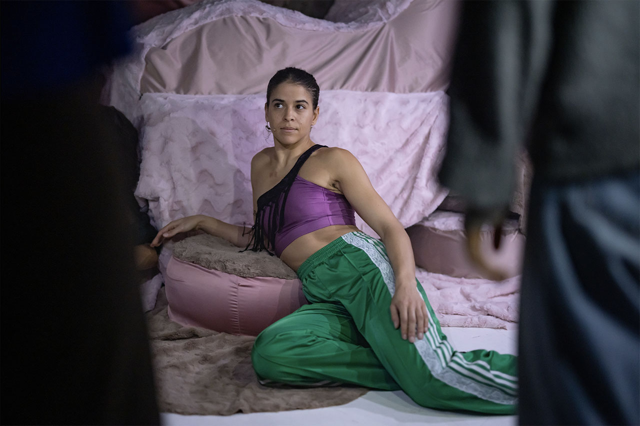 A woman reclines on soft cushions, glancing towards two unseen figures. The scene feels tense and intimate.