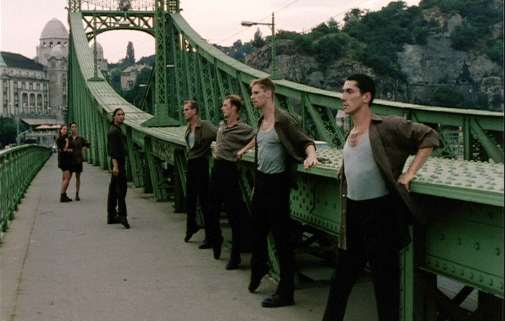 A group of men pause along a striking green steel bridge, framed by historic architecture and rolling hills. The scene captures a quiet yet cinematic urban moment.