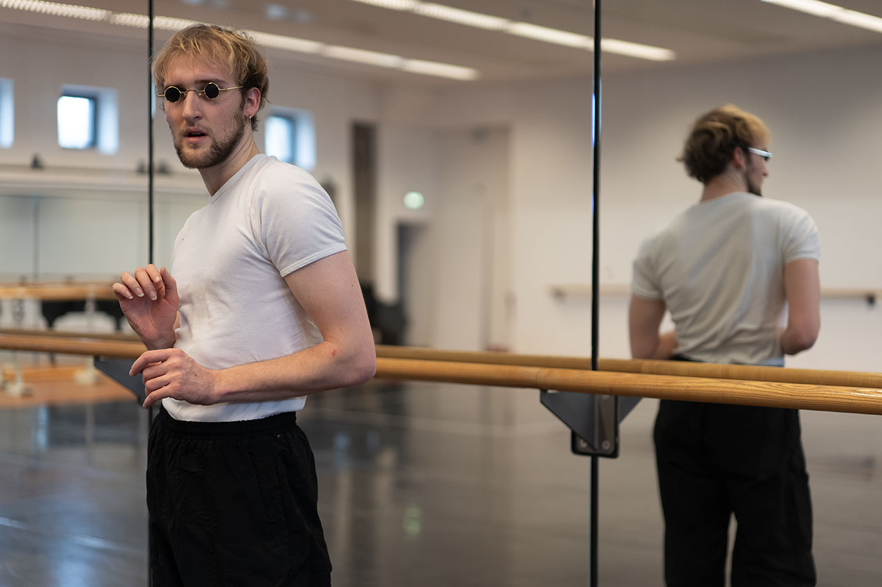 A man pauses beside the mirrored barre in a bright dance studio. His reflection doubles the moment, adding quiet intensity.