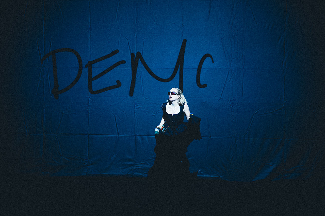 A striking figure stands beneath bold lettering on a deep blue backdrop. The dramatic lighting adds an air of mystery and performance.
