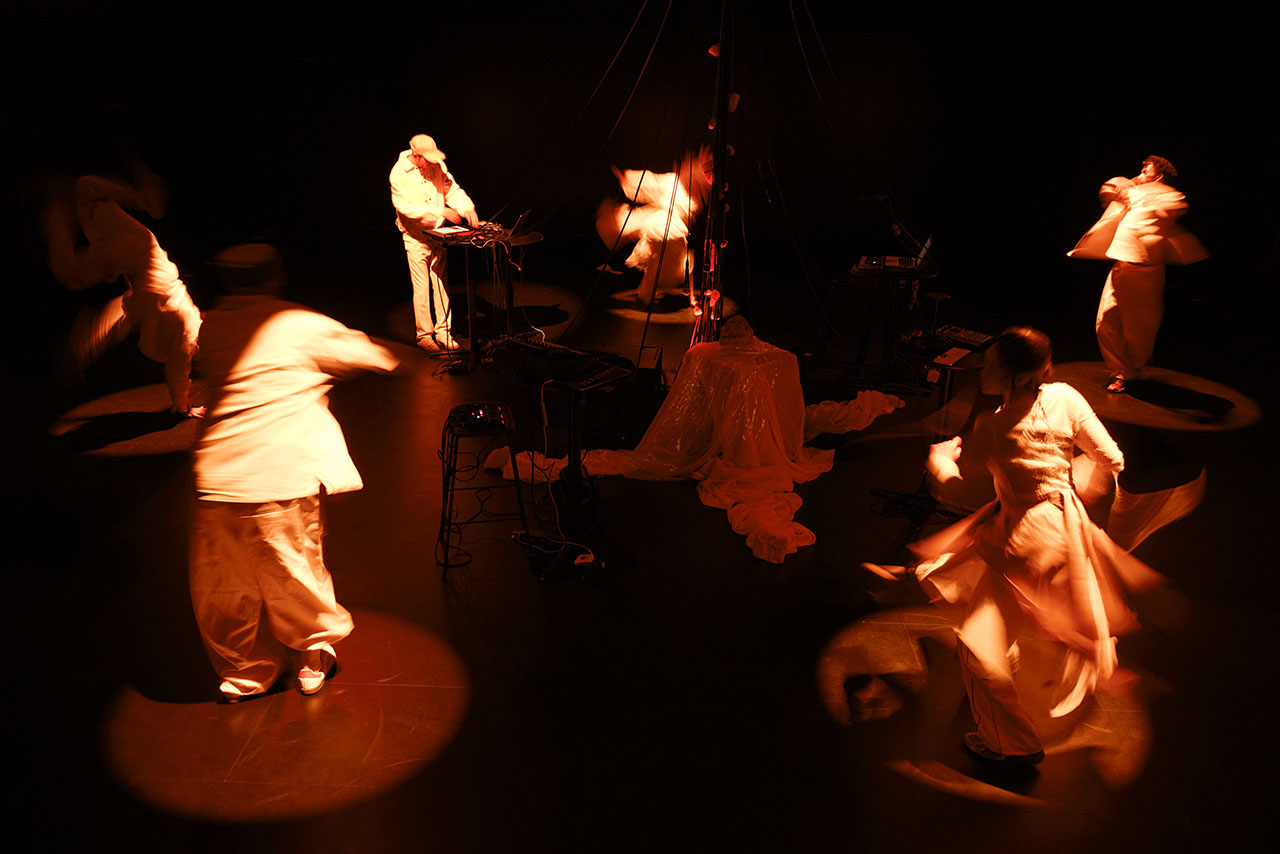 Dancers whirl across a dimly lit stage bathed in deep red light. A live musician stands at the centre, shaping the atmosphere with electronic sound.