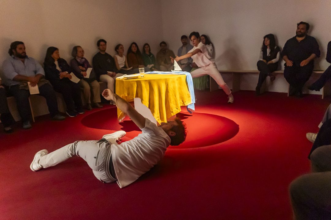 A striking contemporary performance unfolds around a brightly lit table. The audience watches closely as two performers move dynamically across the red floor.
