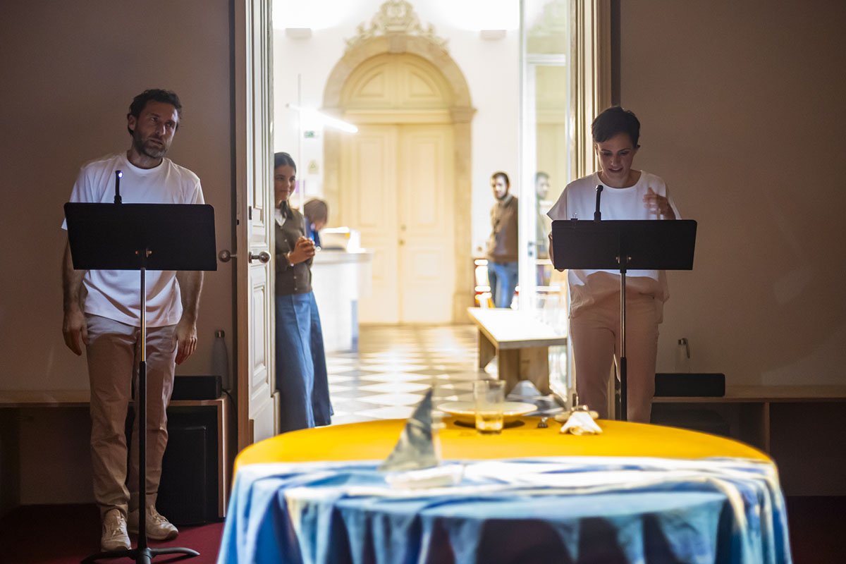Two speakers present a reading in a softly lit gallery setting. Onlookers watch from the adjoining room.