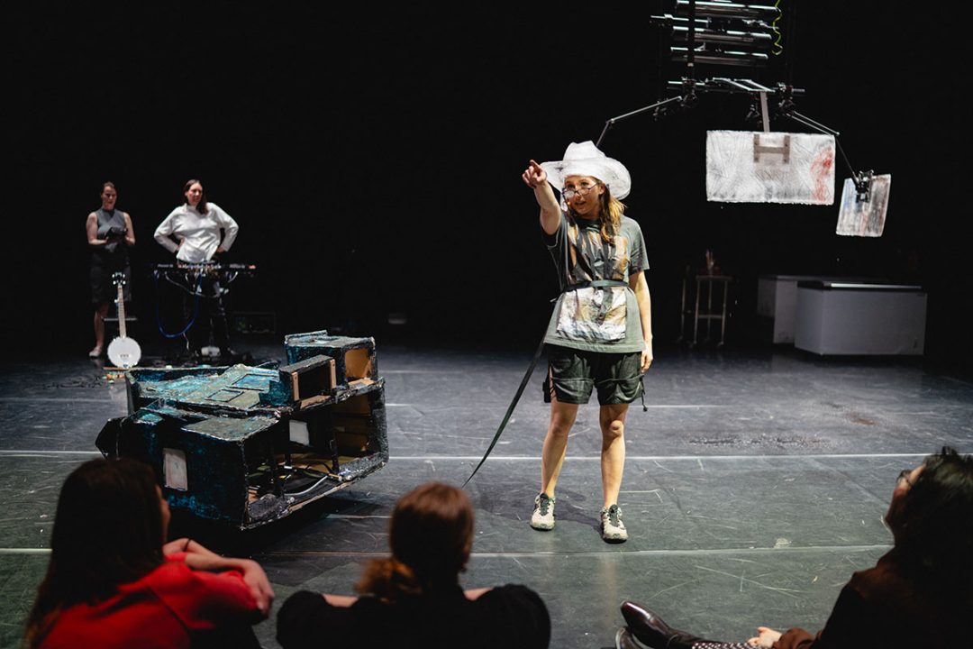 A dynamic moment from an experimental theatre performance. The lead performer gestures towards the audience as musicians stand in the background.
