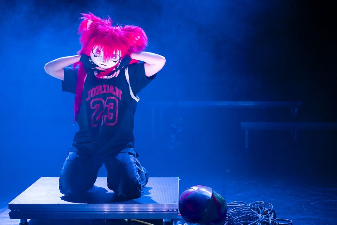 Performer in pink wig kneeling under blue stage lights