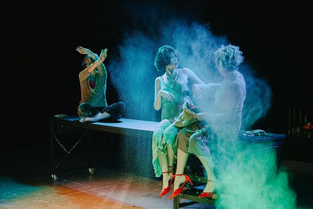 Three performers amid coloured powder on darkened stage