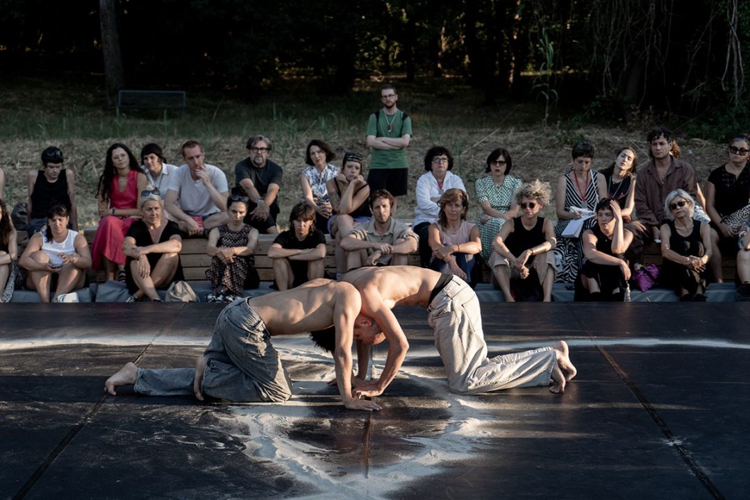 Two shirtless dancers kneeling before seated audience outdoors