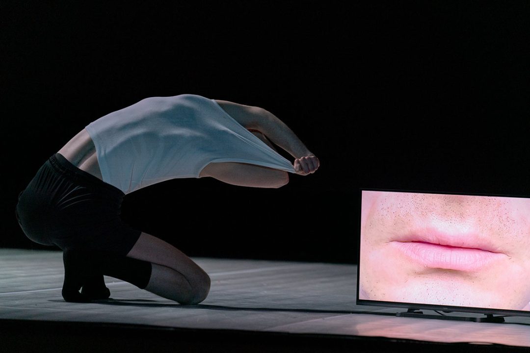 Performer kneeling, pulling shirt, screen shows close-up lips