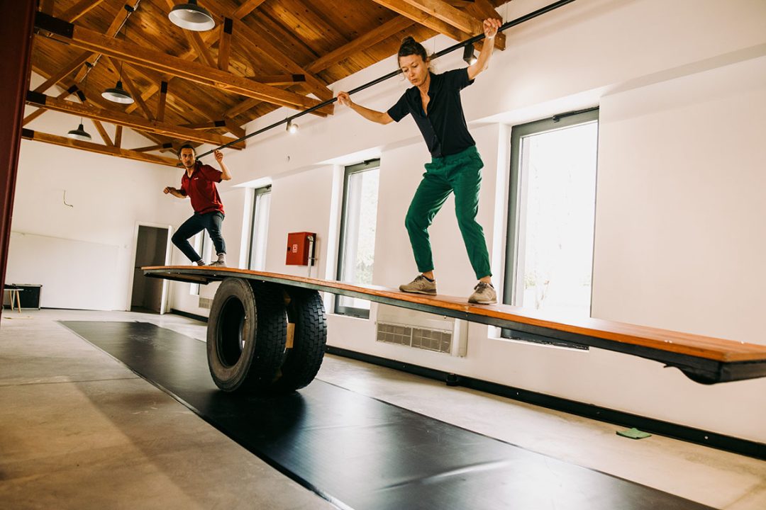 Two people balancing on plank over tyres indoors
