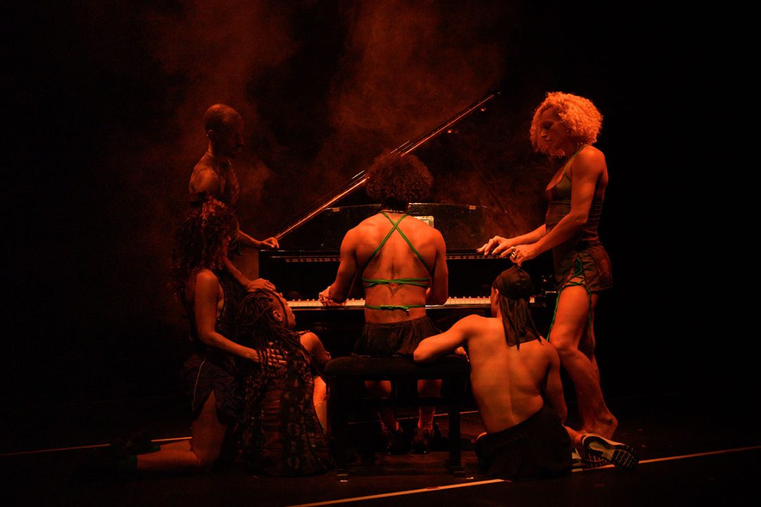 Performers surround a grand piano under dramatic red stage lighting.