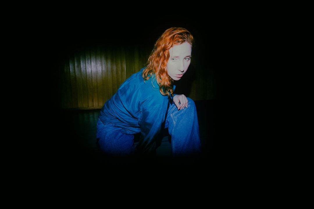 Red-haired woman crouching in blue clothing against dark background