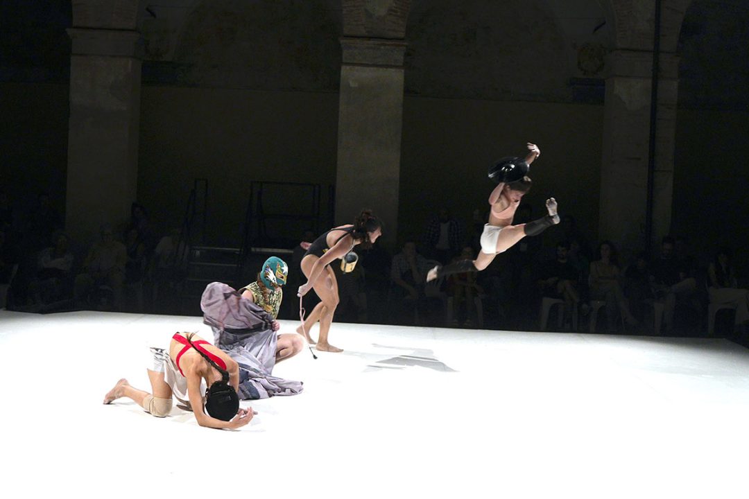 Masked performers dance in a brightly lit theatre ring
