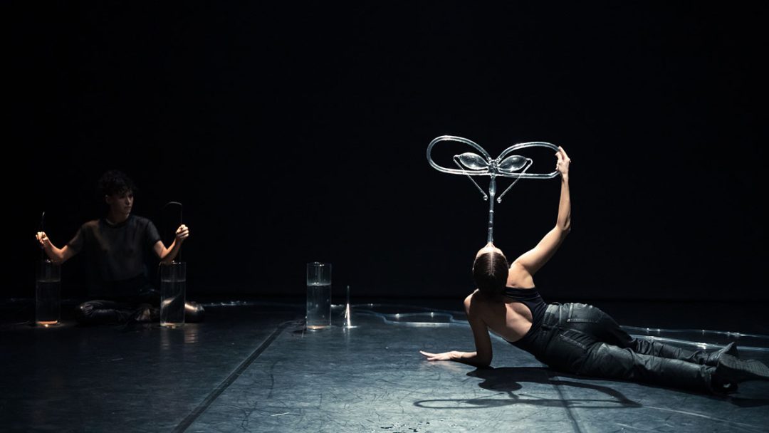 Performer balances looped metal sculpture in dark theatre